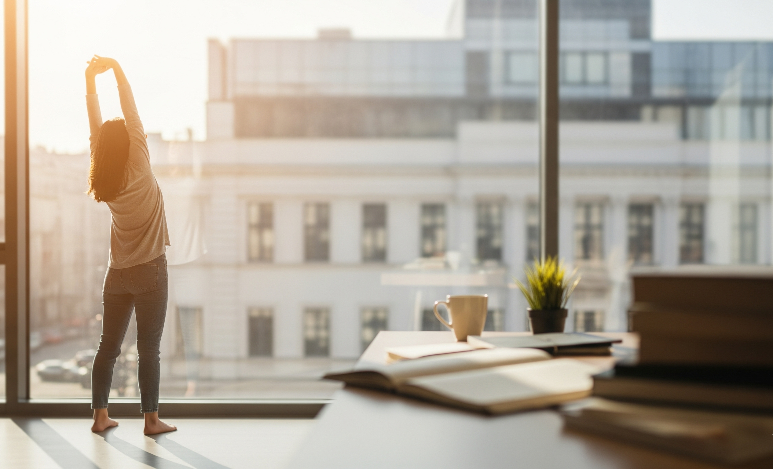 Pessoa se alongando e espreguiçando perto de uma janela com vista urbana, em uma pausa produtiva dos estudos, com uma mesa com livros e xícara de café em primeiro plano.